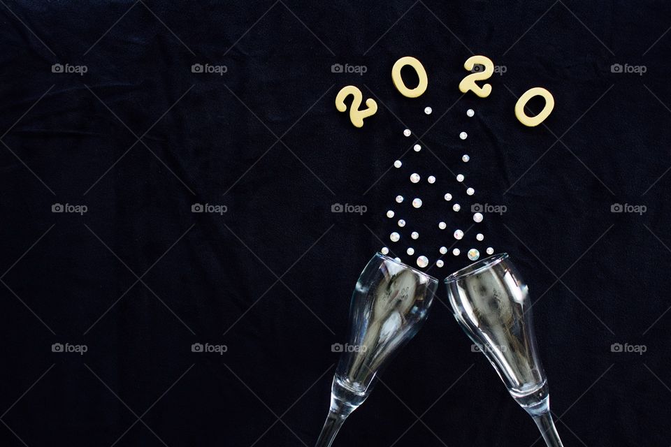 Flat lay of the numbers ‘2020’ in gold above gleaming champagne glasses with rhinestone bubbles on black background - copy space on left side of landscape format