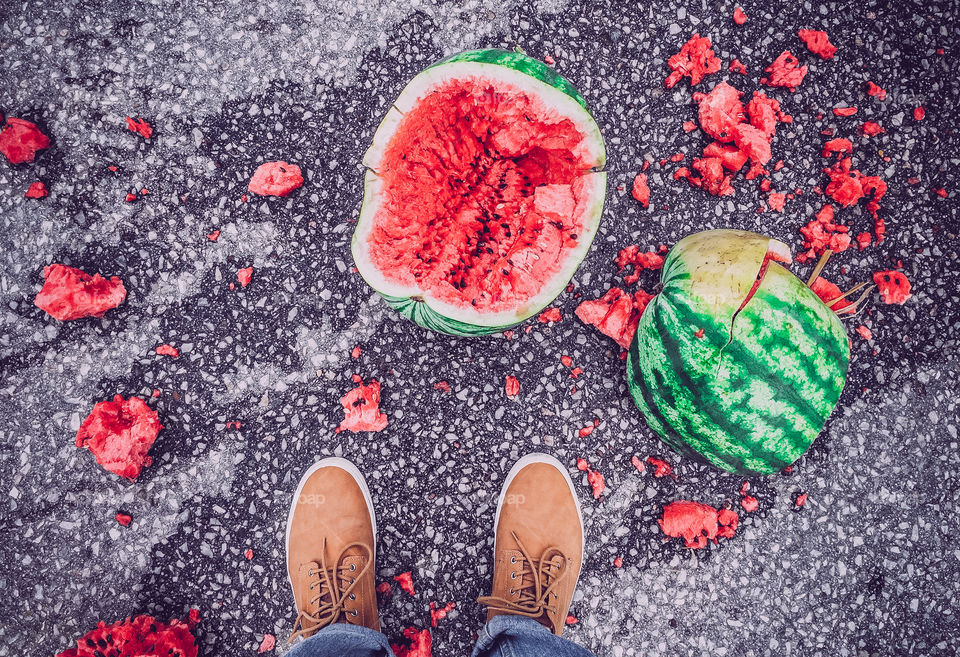 Top view of a smashed watermelon on the ground in front of the feet.