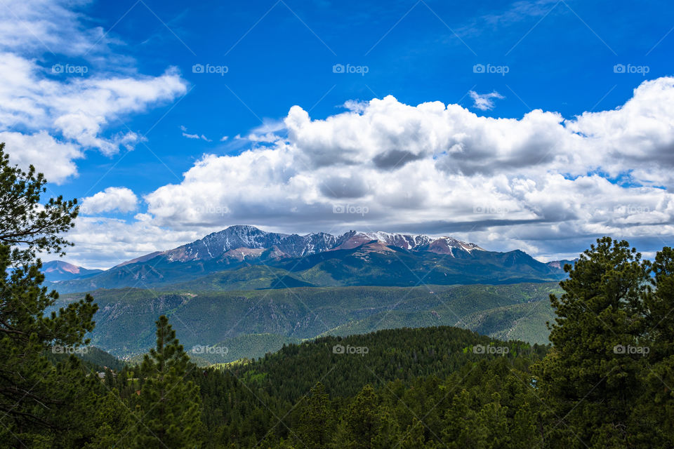Pikes Peak in the Colorado Rocky Mountains sits beneath a sky of rich blue and puffy white. Summer sun streaks the Fourteener's slopes, along with the remnants of the winter's snow.