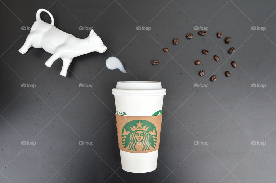 Starbucks coffee cup laid out on a black table with coffee beans appearing to sprout out of the top and milk poured from a ceramic cow creamer