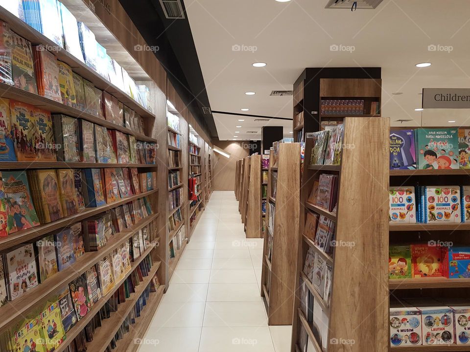 Hundreds of book on wooden shelves, books neatly arranged, long shelves in a bookstore, various books avaliable, shelves made of wood, white floor, bright room light, education book