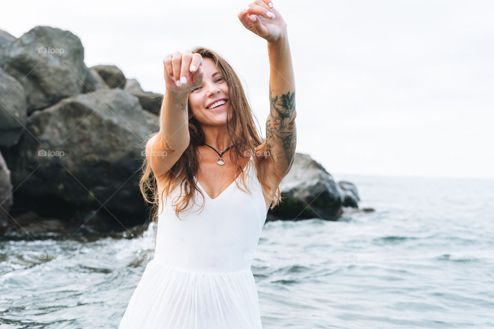 Young carefree woman with long hair in white dress enjoying life on sea beach, people from behind