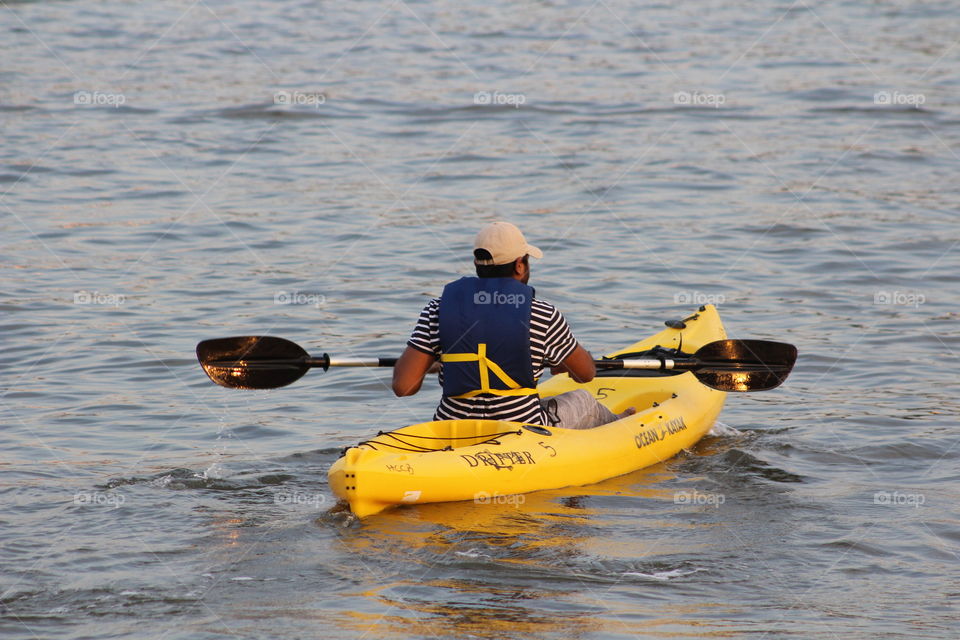 Man in blue life vest, striped shirt and baseball cap paddles bright yellow canoe on Hudson River on May evening