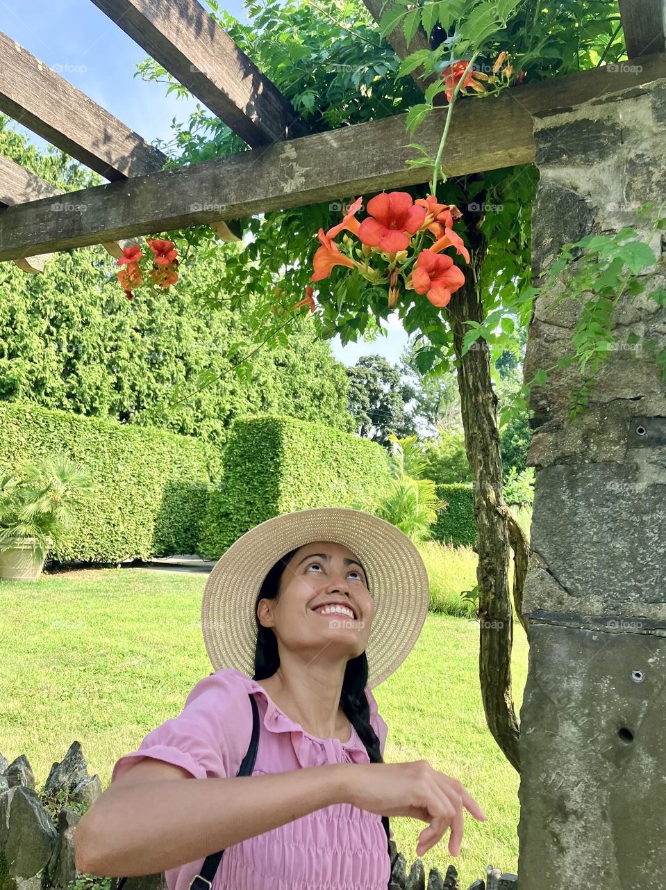 An upbeat woman looking up to trumpet vine red flowers wearing light yellow hat and a pink dress in a sunny day.