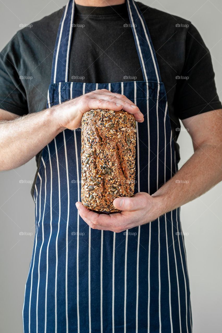 Close-up at bakers hands holding a loaf of whole grain, multi seeded bread in front of him.