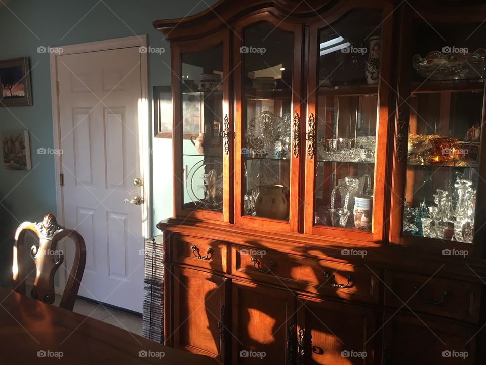 A china cabinet light up by light from a window, showing off the warm brown wood and the fine china inside. 