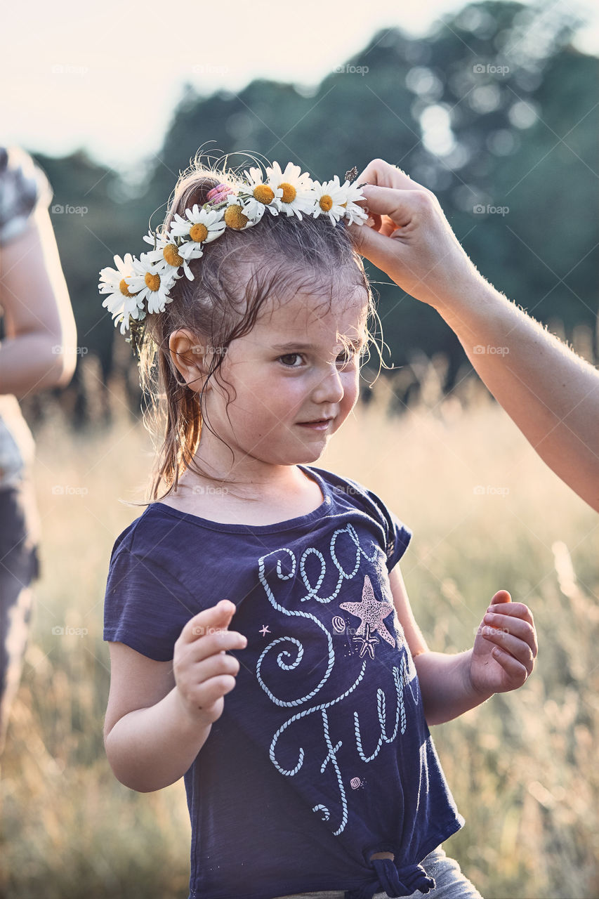 Mother putting a coronet of wild flowers on a head of little girl. Family spending time together on a meadow, close to nature. Parents and children playing together. Candid people, real moments, authentic situations